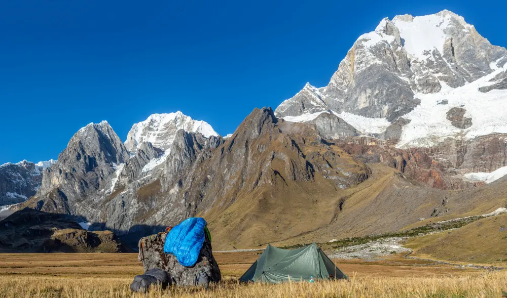 landscape-shot-of-a-green-tent-near-a-rock-in-a-field-surrounded-by-mountains-covered-in-snow